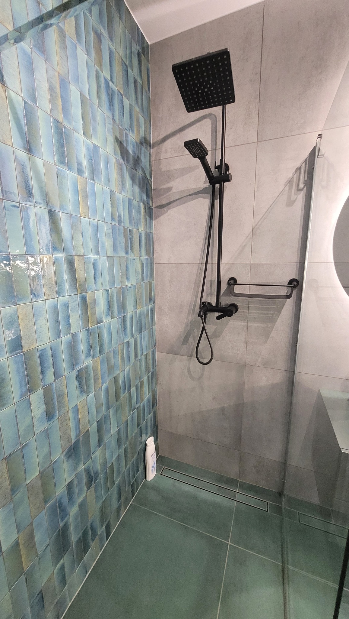 Nowoczesna kabina prysznicowa z deszczownicą w kolorze czarnym, wyłożona turkusowymi płytkami o nieregularnej fakturze, z odpływem liniowym w podłodze i butelką kosmetyku przy ścianie.