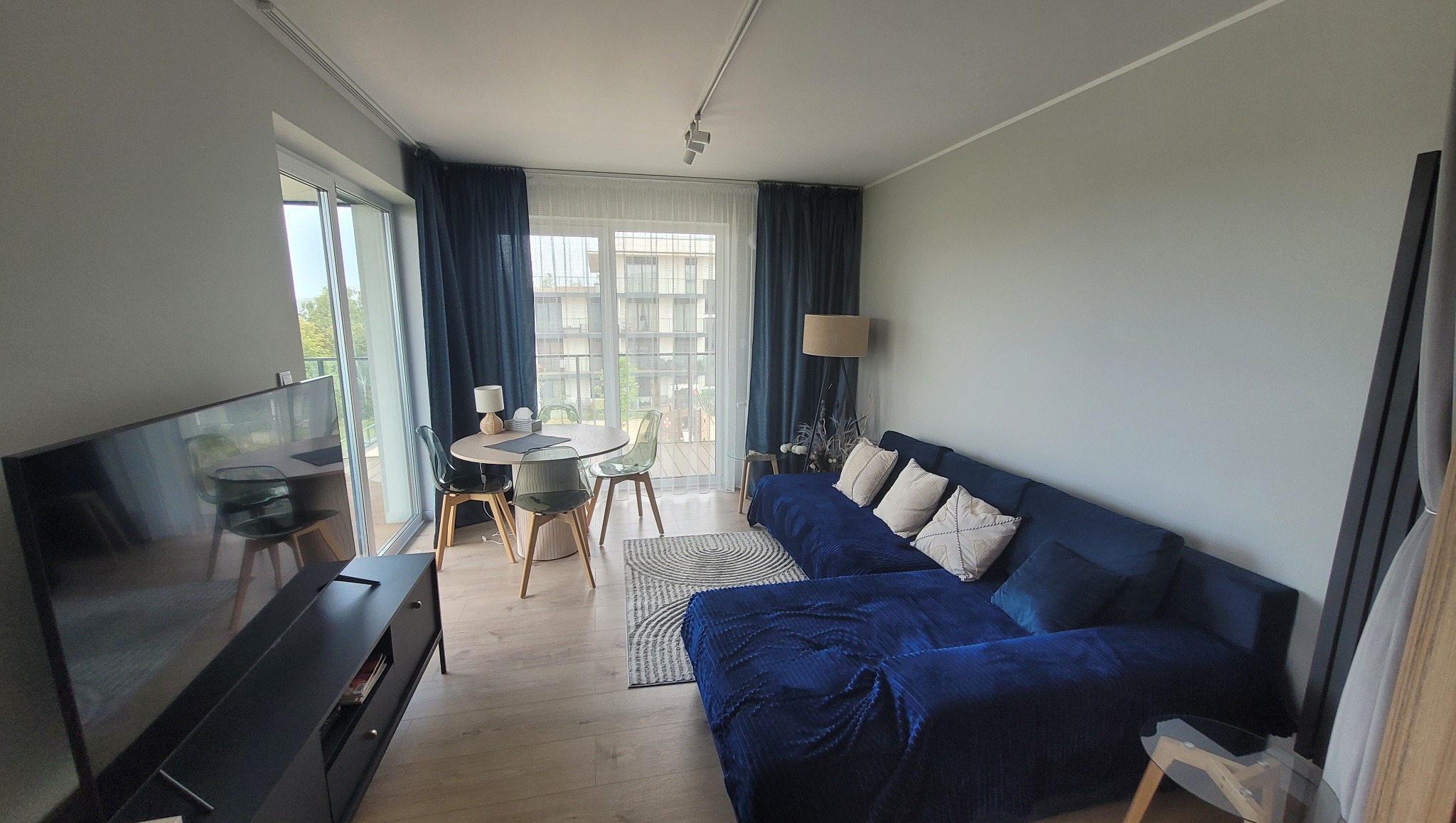 Jasne wnętrze salonu z niebieską sofą narożną, okrągłym stołem z transparentnymi krzesłami i dużym oknem balkonowym z ciemnymi zasłonami.