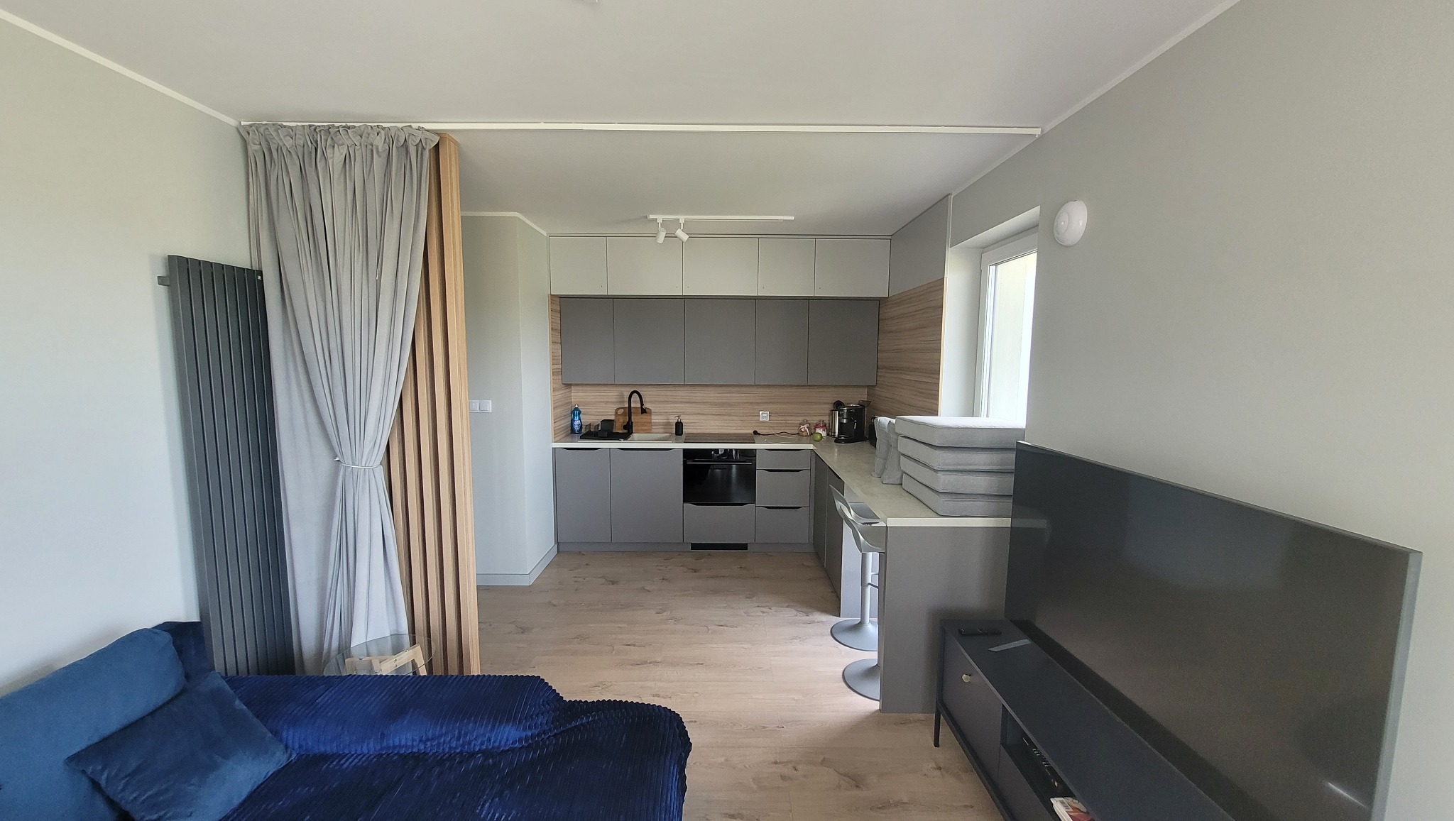 Nowoczesne wnętrze mieszkania: szara kuchnia z drewnianym panelem dekoracyjnym, zasłona oddzielająca przestrzeń, niebieska sofa i telewizor.