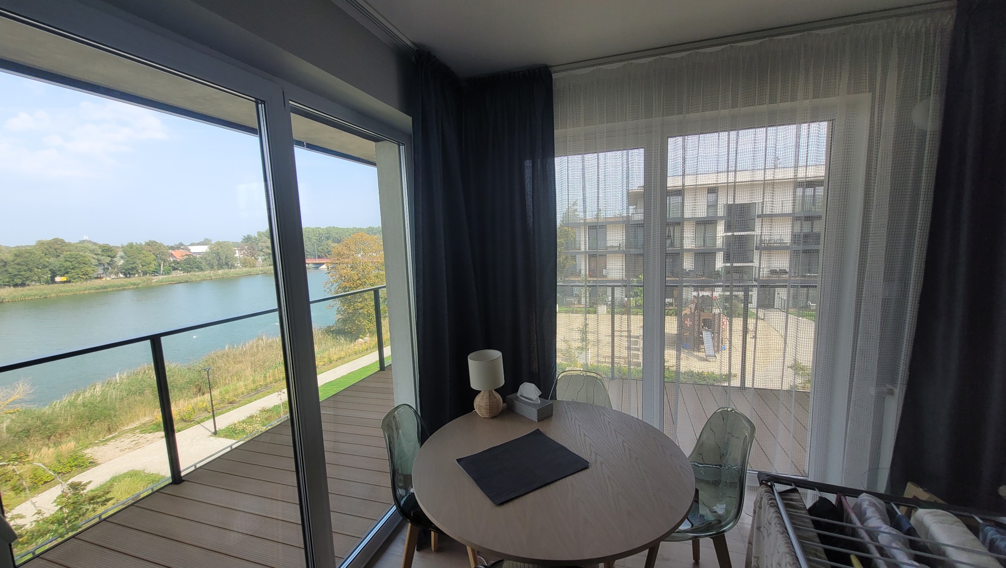 Jasne wnętrze z okrągłym stołem, krzesłami z tworzywa i widokiem na jezioro z balkonu, zasłonięte częściowo czarnymi i białymi zasłonami.