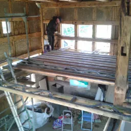 Wnętrze domu w trakcie ocieplania, widoczna drewniana konstrukcja ścian, ocieplenie pomiędzy belkami, pracownik w kombinezonie przy oknie, drabiny i materiały budowlane.