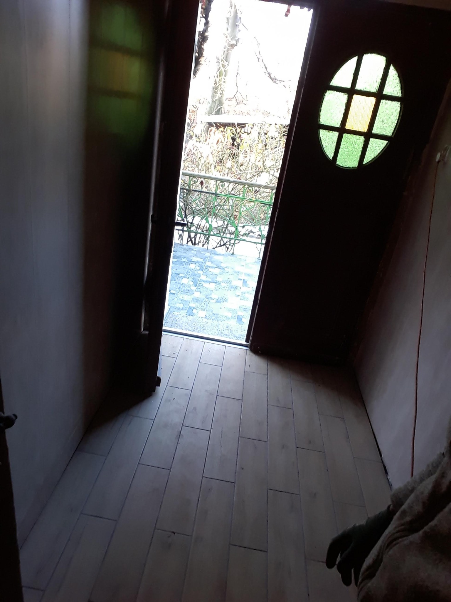 Podłoga wyłożona płytkami imitującymi drewno w pomieszczeniu z oknem w kształcie elipsy z kolorowymi szybkami, widok na balkon i drzewa.