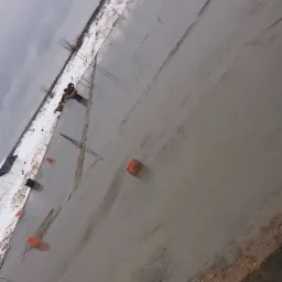 Świeżo wylany beton na fundamencie z widocznym zbrojeniem i pomarańczowymi ogranicznikami, śnieg w tle.