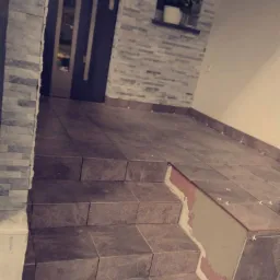 Układanie płytek na schodach wejściowych do domu; widoczne krzyżyki dystansowe, częściowo wykończona ściana z kamienia dekoracyjnego i okno z czerwonymi kwiatami.