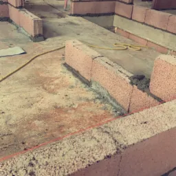 Murowanie ścian z czerwonych bloczków w surowym wnętrzu poddasza, widoczne narzędzia murarskie i elementy konstrukcji dachu.