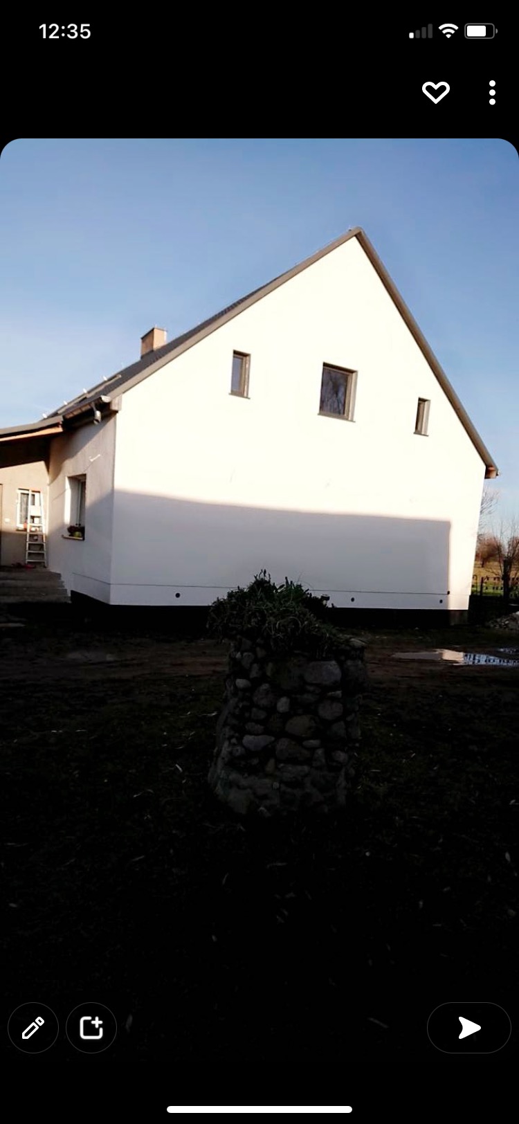 Biały dom jednorodzinny z ocieploną elewacją, widoczne okna, fragment dachu i drabina oparta o ścianę, na pierwszym planie kamienna studzienka z roślinami.