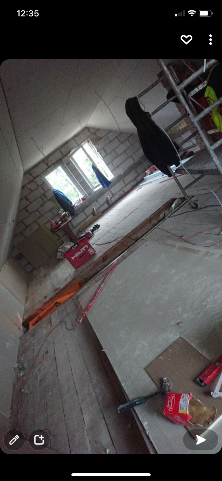 Wnętrze poddasza w trakcie prac: surowe, murowane ściany z widocznymi oknami, nieukończona podłoga, narzędzia budowlane i drabina rusztowania.