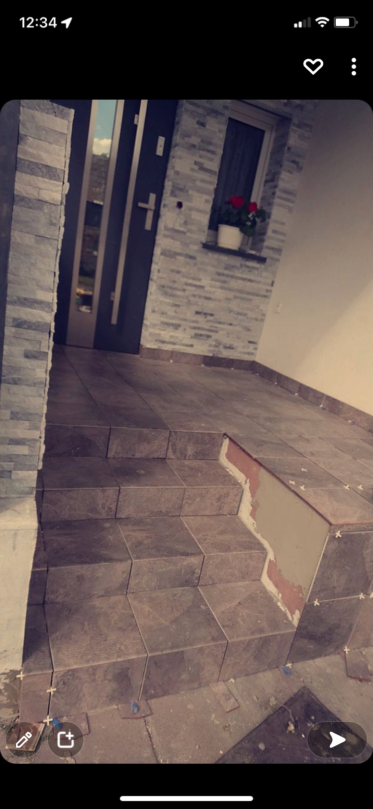 Układanie płytek na schodach wejściowych do domu; widoczne krzyżyki dystansowe, częściowo wykończona ściana z kamienia dekoracyjnego i okno z czerwonymi kwiatami.