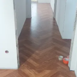 Ułożona podłoga w jodełkę z brązowych paneli, widoczne taśmy malarskie przy progu i surowe wykończenie ścian w tle.