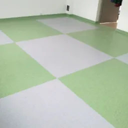 Szachownica z jasnozielonych i szarych kwadratowych płytek podłogowych w pustym pomieszczeniu z białymi ścianami i widocznymi listwami przypodłogowymi w kolorze zielonym.