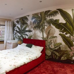 Pictograf - Sypialnia z czerwonym łóżkiem i fototapetą przedstawiającą tropikalną roślinność, widoczny również łapacz snów przy oknie.