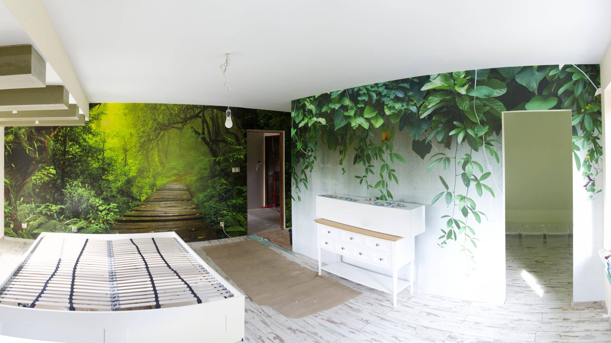 Pokój z fototapetami: leśna ścieżka na jednej ścianie, motyw roślinny na drugiej, w tle białe meble i niedokończone łóżko.