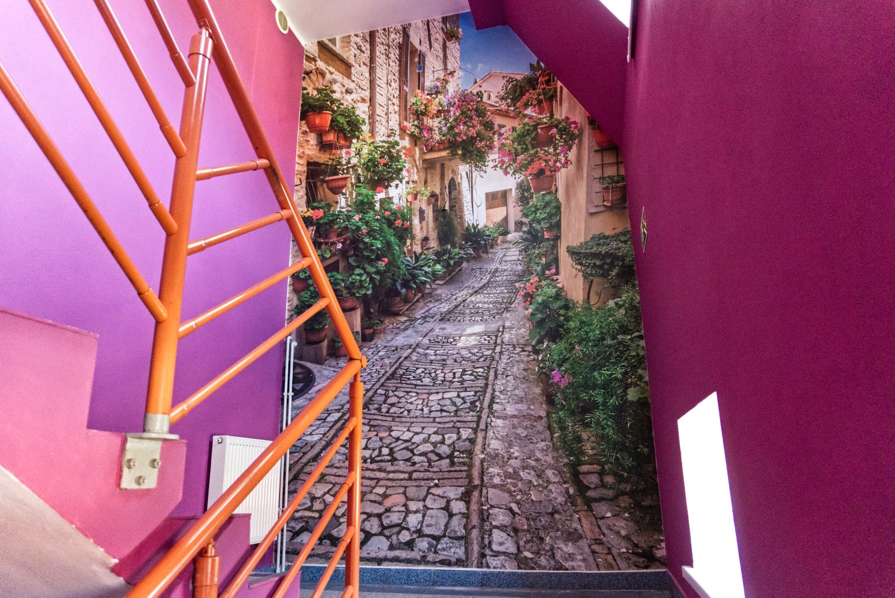 Fototapeta z motywem włoskiej uliczki z kwiatami na ścianie klatki schodowej w Środzie Śląskiej, widoczna pomarańczowa barierka schodów.