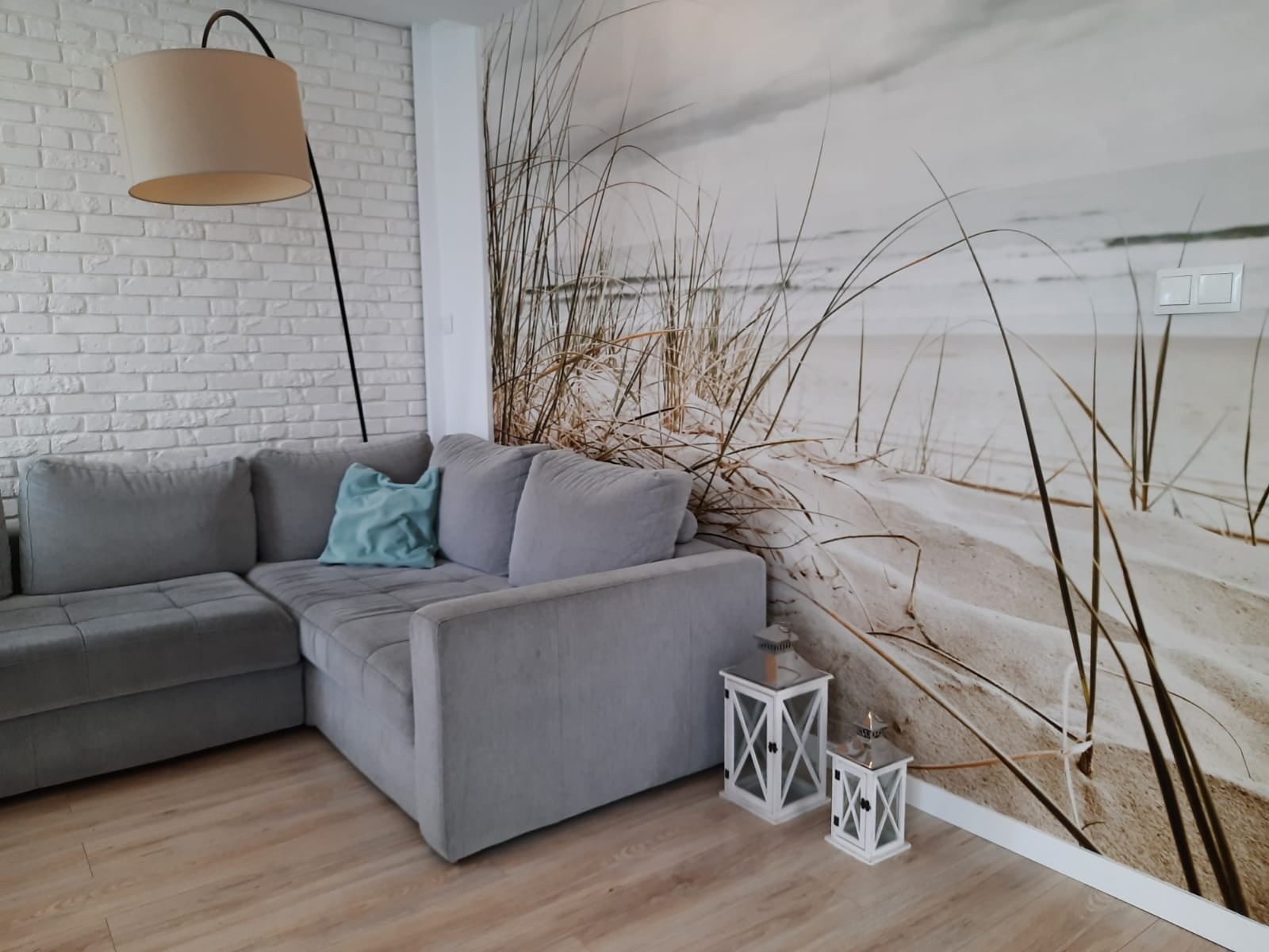 Szary narożnik z turkusową poduszką na tle fototapety z motywem plaży i wydm, biała ściana z cegły i lampa łukowa.