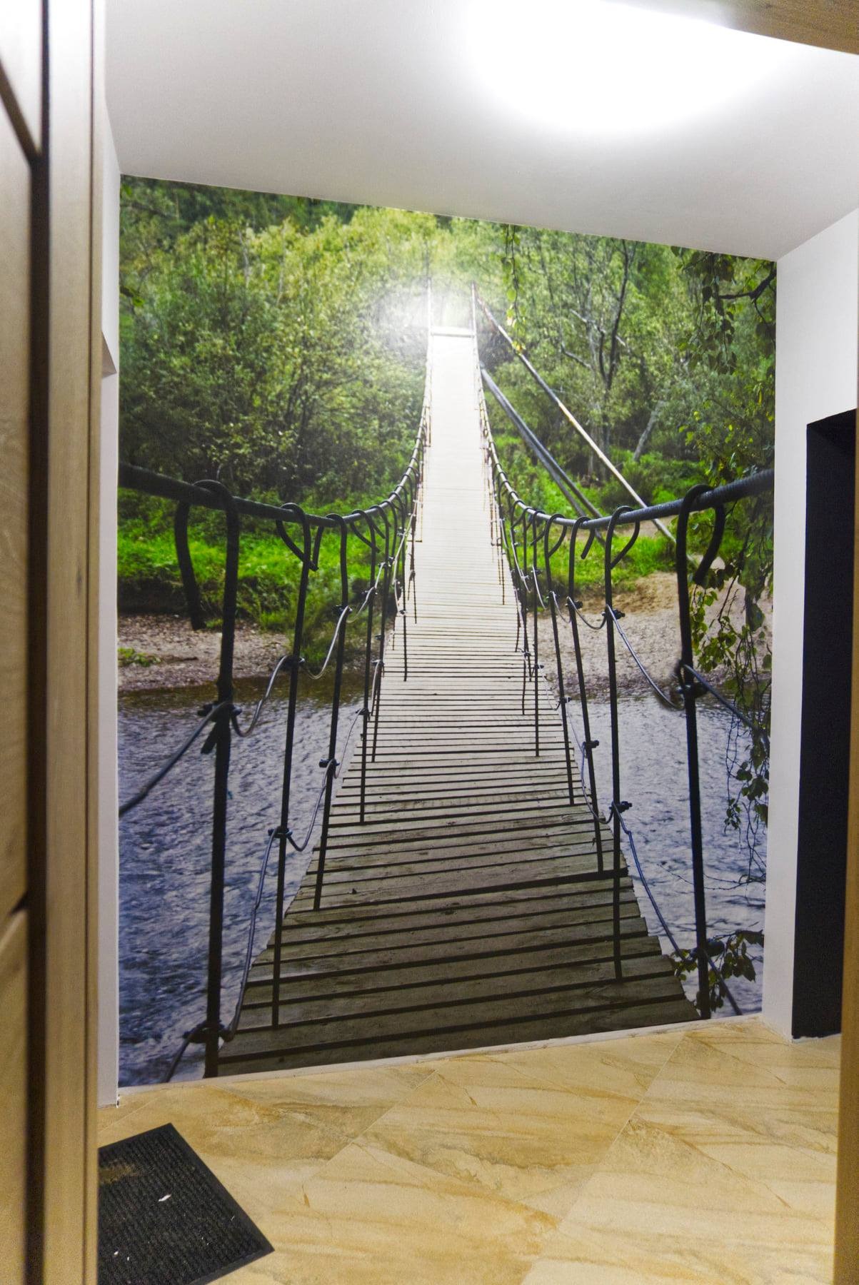 Fototapeta na ścianie przedstawiająca most wiszący nad rzeką, z widocznymi drzewami w tle, oświetlona punktowo, z fragmentem parkietu na podłodze.