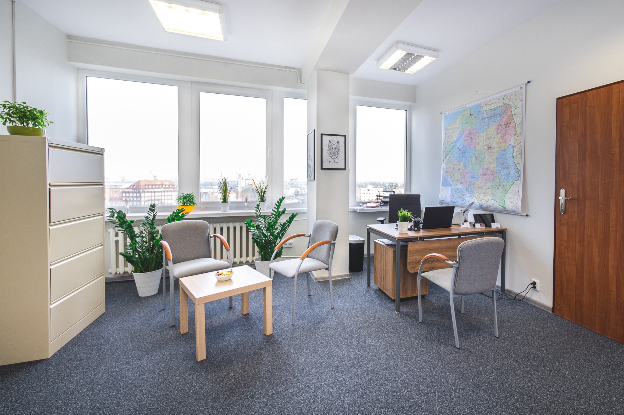 Jasne wnętrze biura z trzema oknami, biurkiem, mapą Polski na ścianie, szafką na dokumenty i stolikiem kawowym z przekąskami