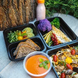 Zestaw posiłków w czarnych pojemnikach na wynos: ryba z grilla, fioletowy kalafior, zielone warzywa, pasta awokado, pieczywo pełnoziarniste, zupa krem pomidorowa z bazylią, sałatka z bakłażanem...
