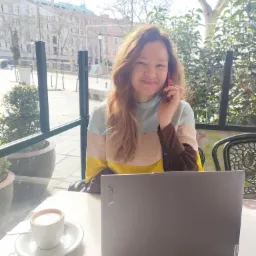 Kobieta z laptopem i kawą w kawiarni, rozmawia przez telefon. W tle budynki miejskie w słoneczny dzień. Nauka języka hiszpańskiego online.