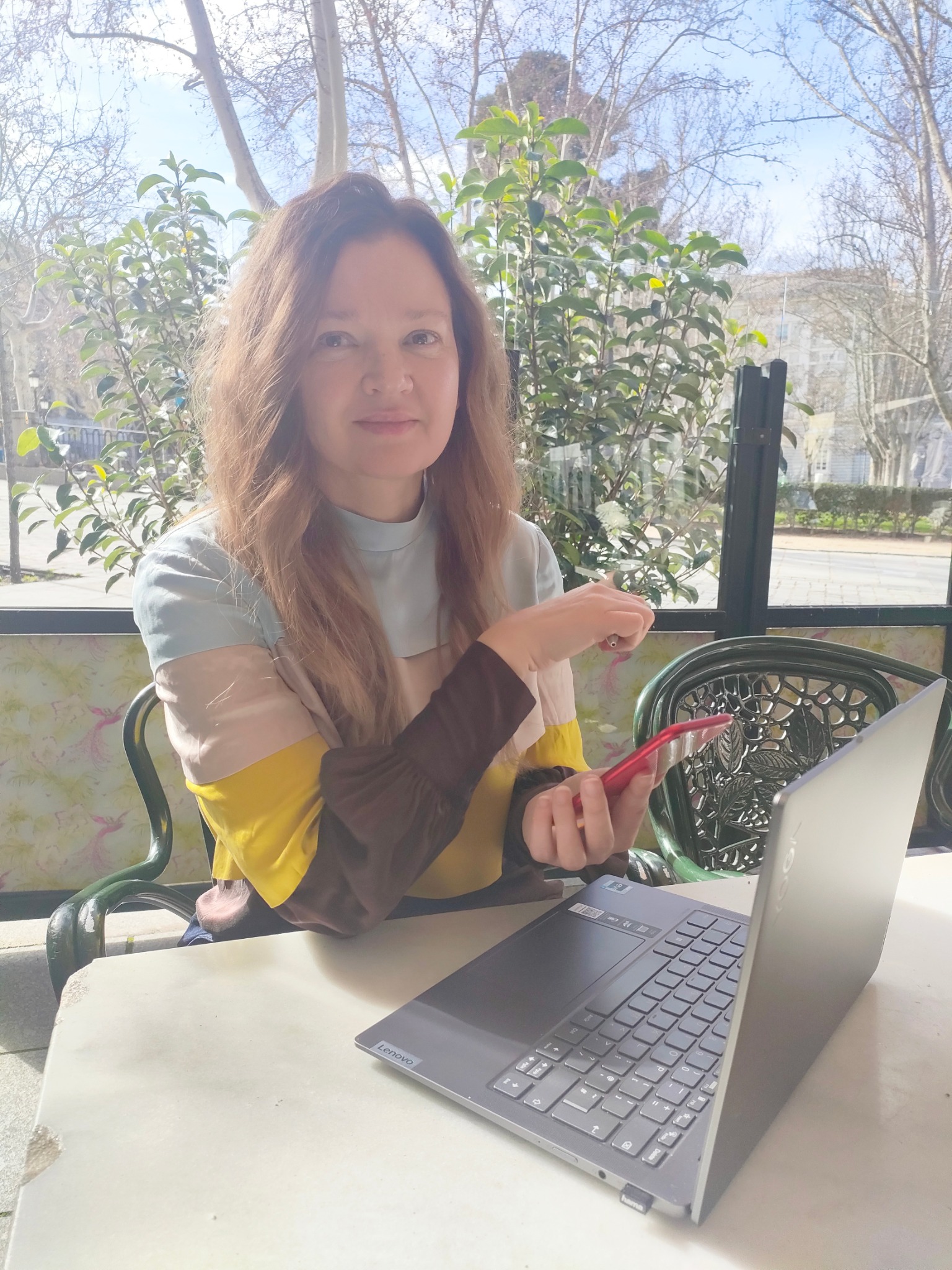 Kobieta z laptopem i telefonem siedzi na zewnątrz, prawdopodobnie ucząc się lub pracując zdalnie. W tle zieleń i budynki, sugerujące miejską scenerię.