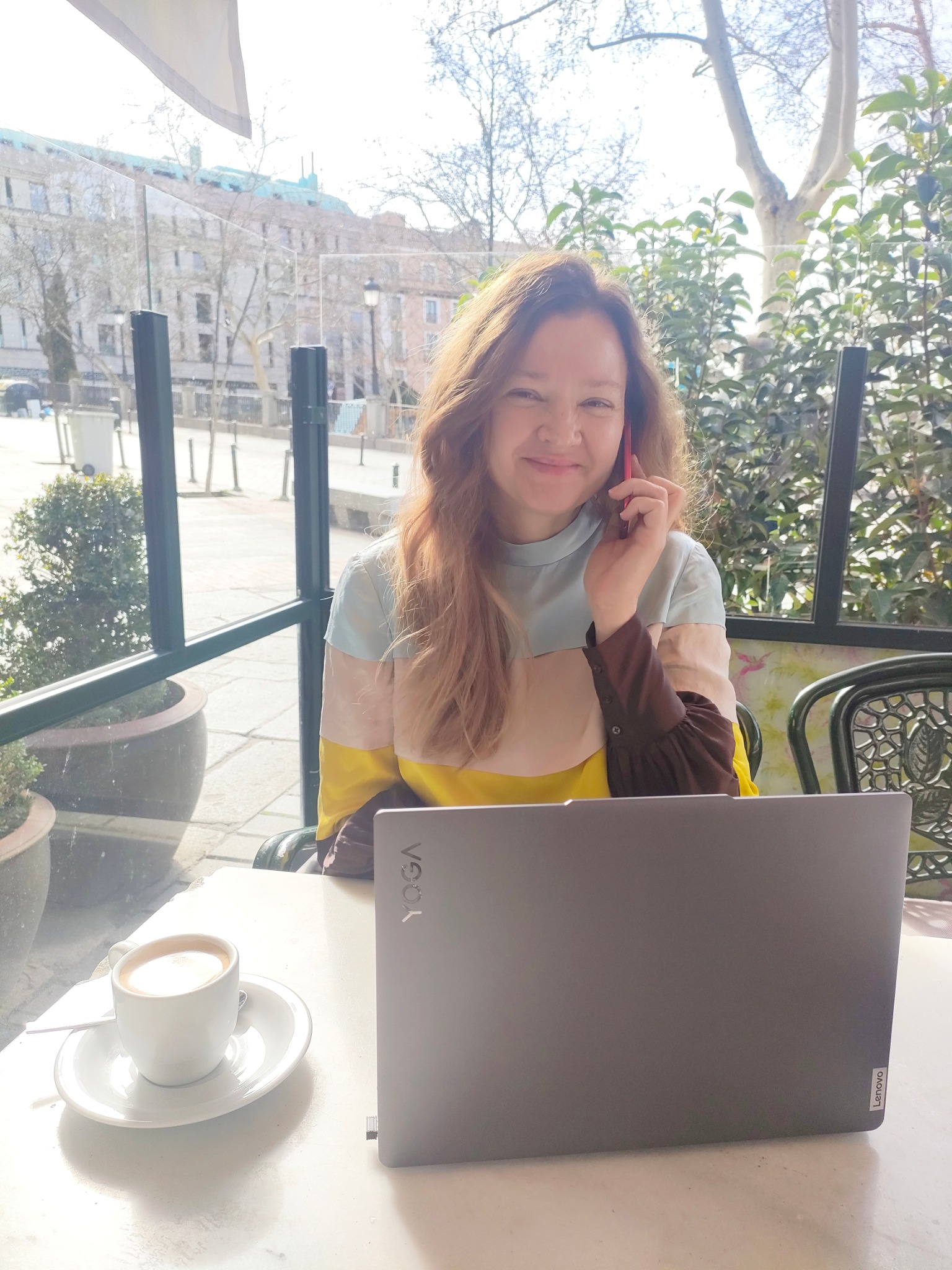 Kobieta z laptopem i kawą w kawiarni, rozmawia przez telefon. W tle budynki miejskie w słoneczny dzień. Nauka języka hiszpańskiego online.