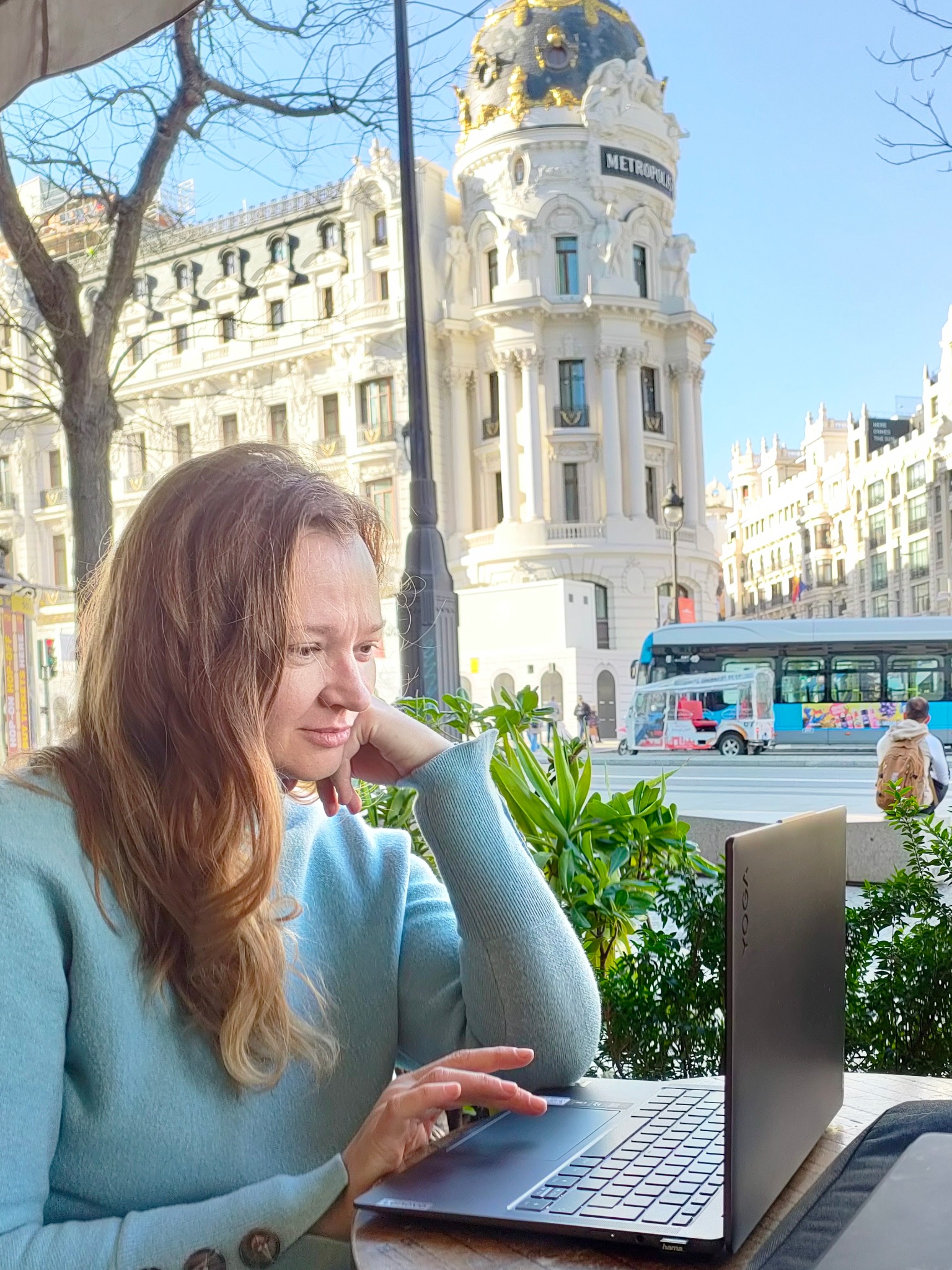 Kobieta z laptopem Yoga w kawiarni na tle architektury Madrytu, być może podczas lekcji online. W tle budynek Metropolis i autobus turystyczny.