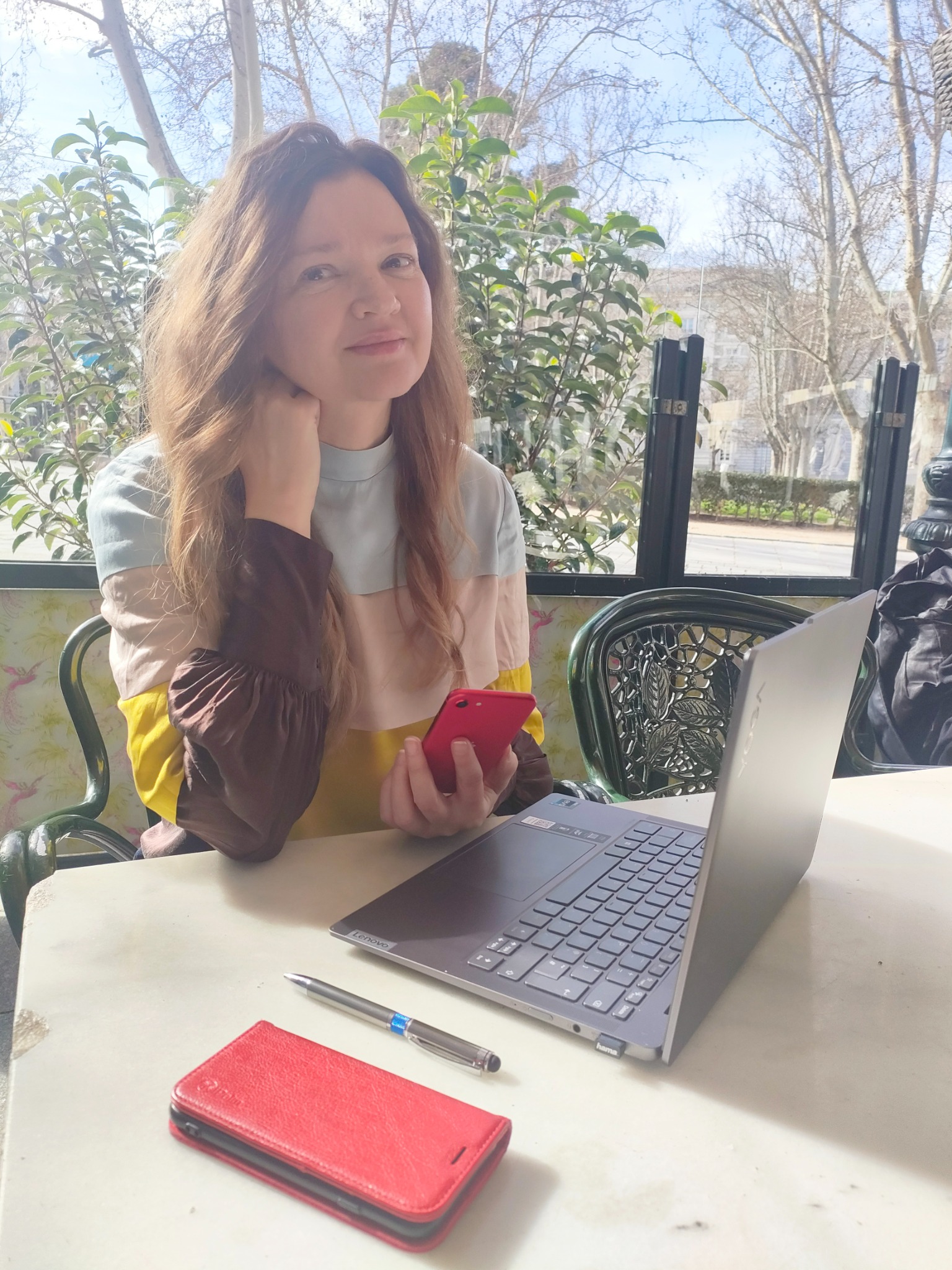 Kobieta z telefonem i laptopem siedzi przy stoliku na zewnątrz, być może podczas nauki języka online. W tle widoczne drzewa i budynki.