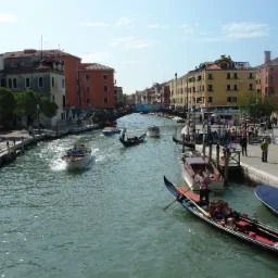 Widok na kanał w Wenecji z gondolami, łodziami motorowymi i pieszym mostem w tle, w słoneczny dzień.