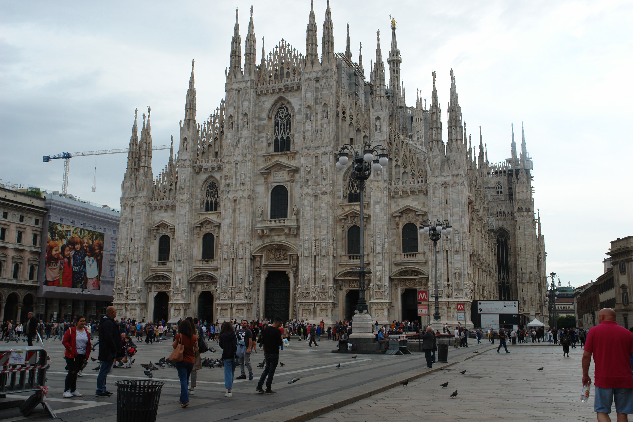 Duomo di Milano, gotycka katedra z białego marmuru, na tle pochmurnego nieba, otoczona placem z przechodniami i gołębiami, z widocznym żurawiem budowlanym w tle.
