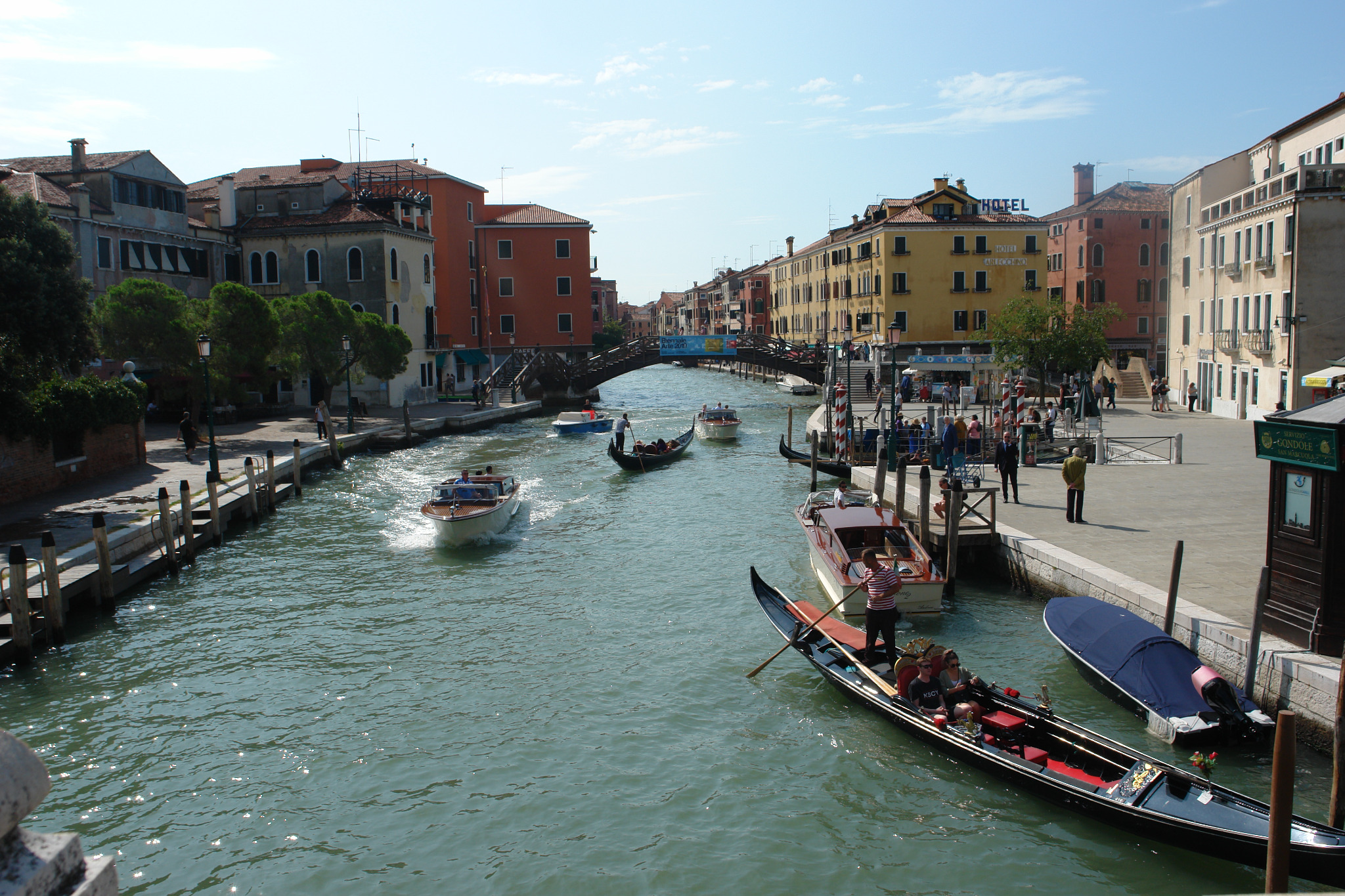 Widok na kanał w Wenecji z gondolami, łodziami motorowymi i pieszym mostem w tle, w słoneczny dzień.
