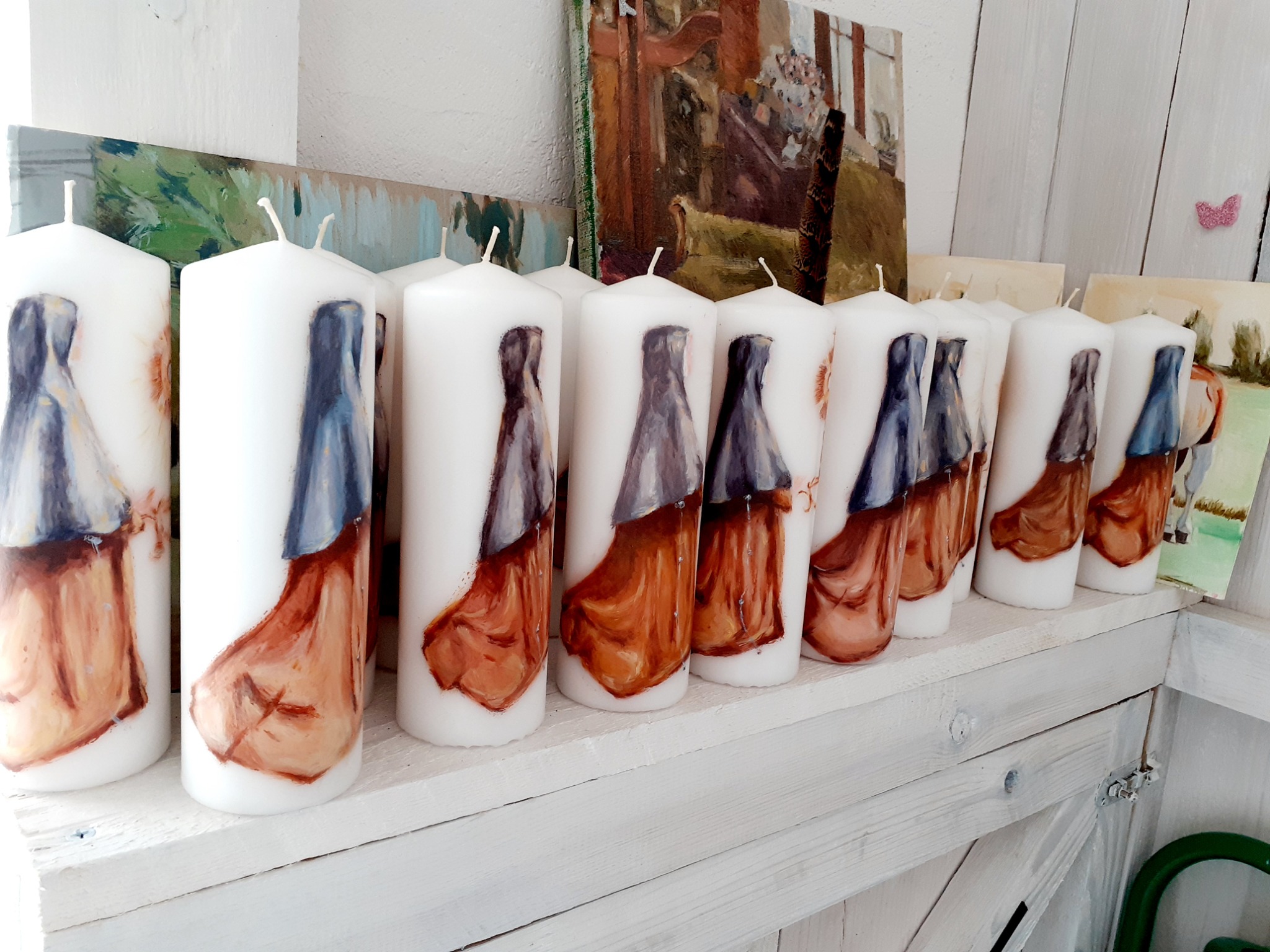 Rząd białych, walcowatych świec z ręcznie malowanymi postaciami w szatach, ustawionych na białej, drewnianej półce, z obrazami w tle.