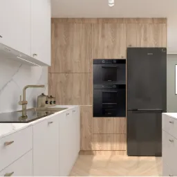 Nowoczesna kuchnia z białymi szafkami z marmurowym blatem, złotą armaturą i drewnianymi frontami szafek górnych, z wbudowanym piekarnikiem i czarną lodówką Samsung po prawej stronie.