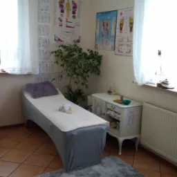 Jasne wnętrze gabinetu z kozetką przykrytą szarą narzutą, fioletową poduszką i stosem białych ręczników, na ścianach wiszą plansze anatomiczne, obok kozetki stoi biała komoda z akcesoriami.