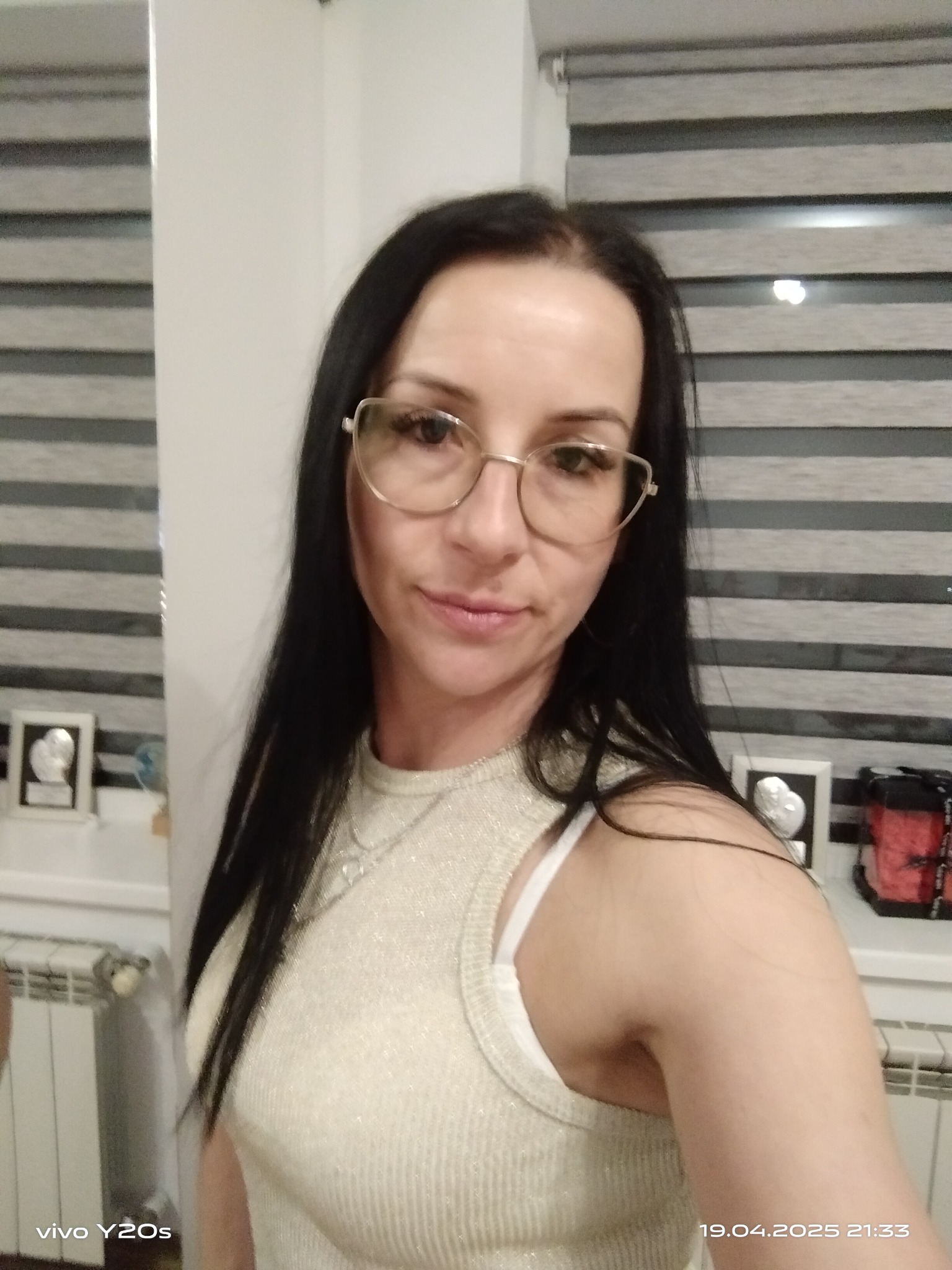 Portret kobiety w okularach, z długimi czarnymi włosami, w jasnej bluzce, na tle żaluzji i dekoracji. Zdjęcie wykonane z bliska, z widocznym datownikiem.