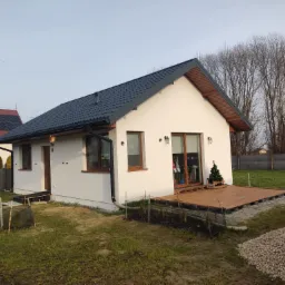 Parterowy dom z białymi ścianami i ciemnym dachem, drewniany taras z małą choinką, widok z ogrodu w pochmurny dzień.