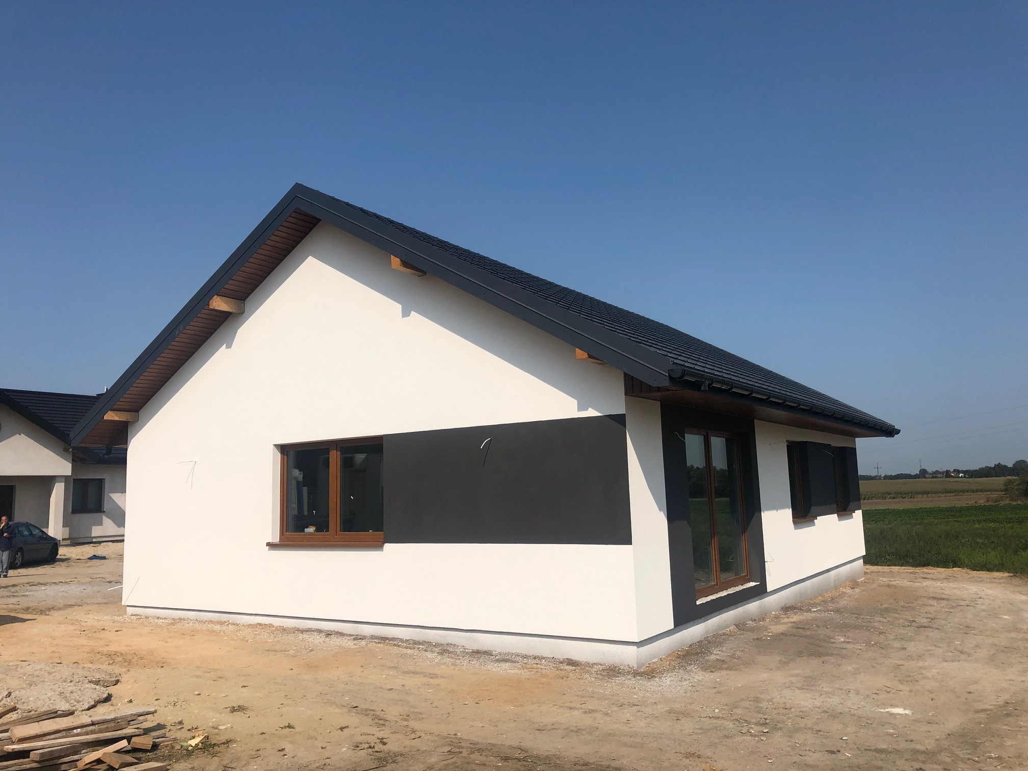 Nowoczesny dom szkieletowy z białymi ścianami i grafitowym pasem elewacyjnym pod oknem, widoczne drewniane ramy okienne i czarna dachówka, w tle pole uprawne.