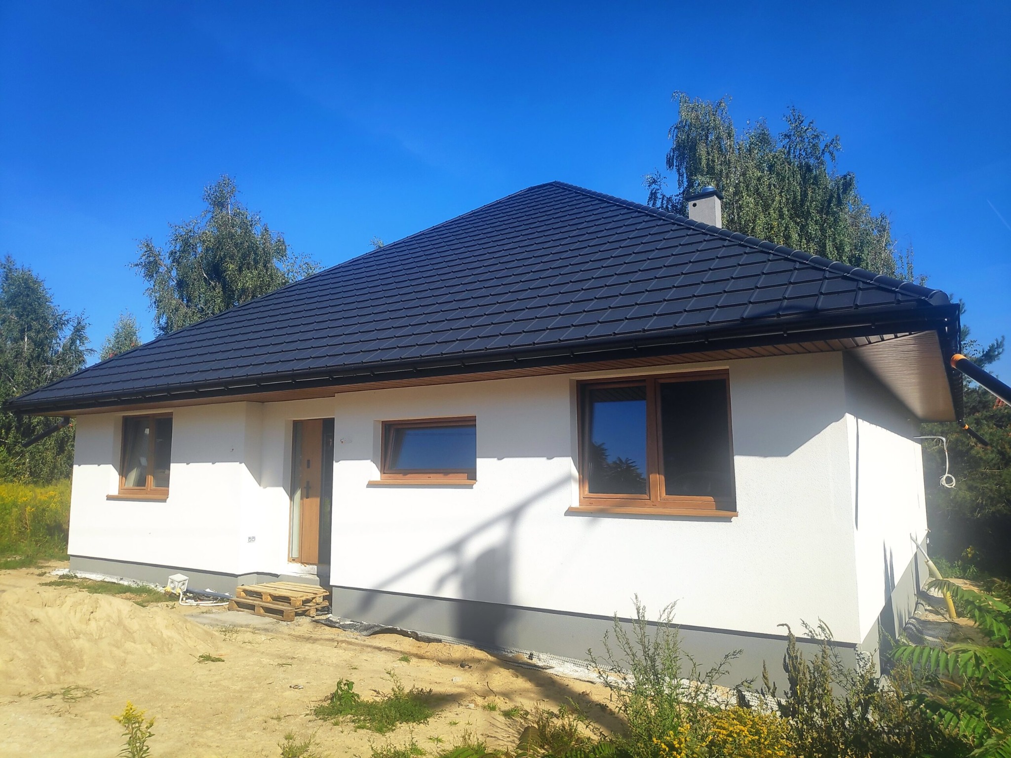 Nowoczesny, parterowy dom z czarnym dachem i jasną elewacją, częściowo wykończony, widoczne palety przed wejściem, błękitne niebo w tle.