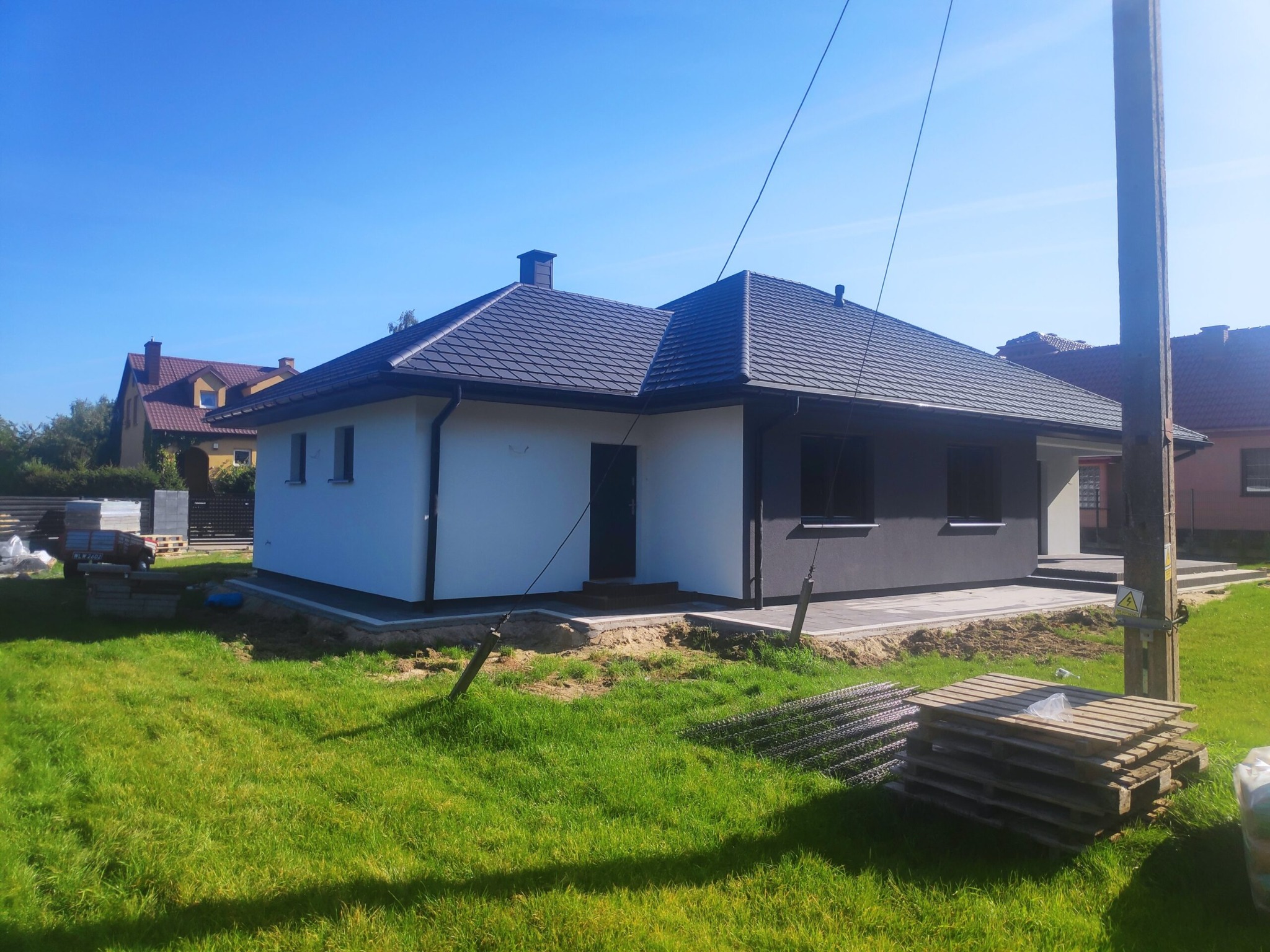 Niedawno wybudowany parterowy dom z dwukolorową elewacją (biała i grafitowa) pod ciemnym dachem, widoczny z boku na tle zielonego trawnika i błękitnego nieba.