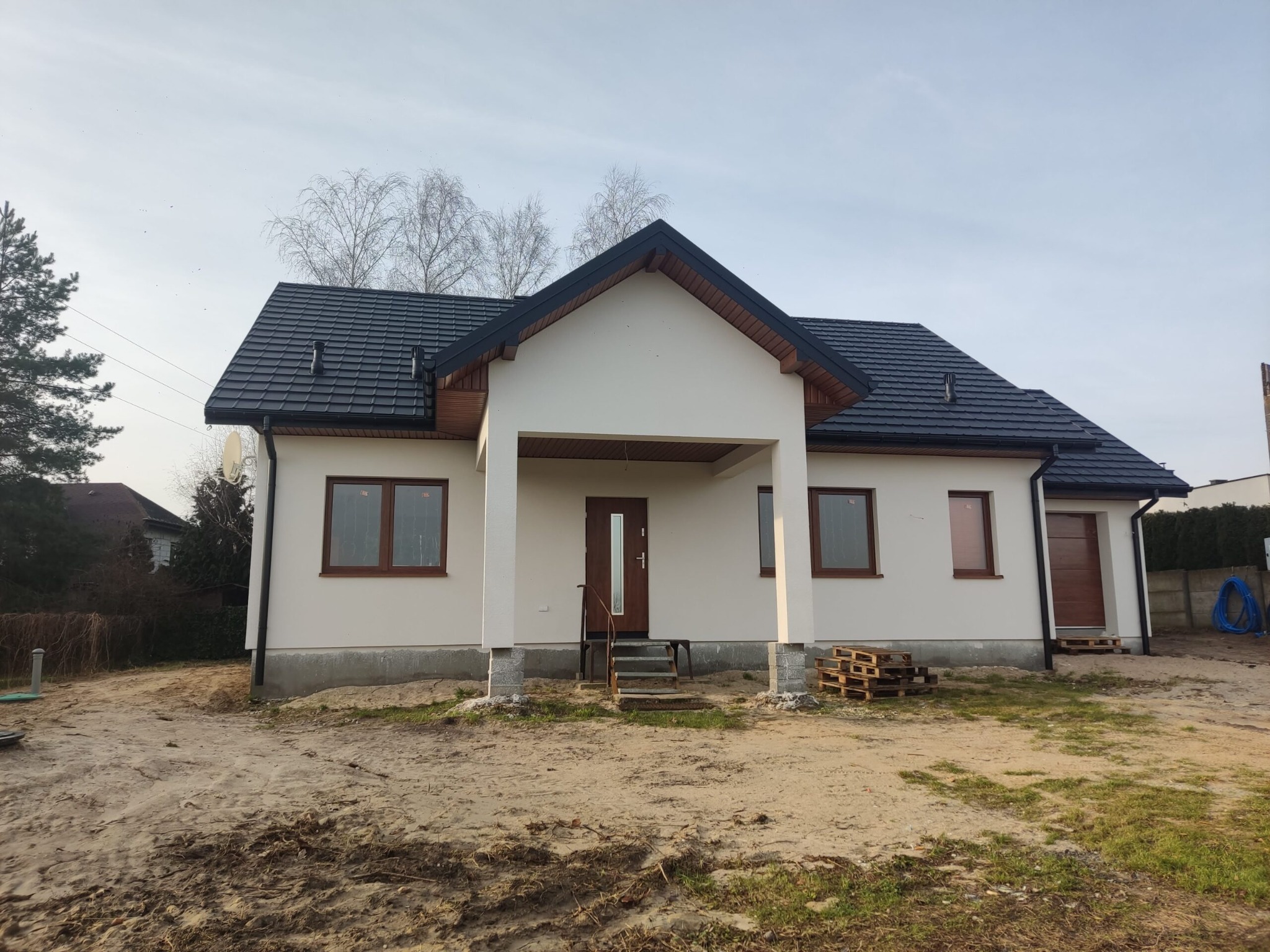 Niedawno wybudowany, parterowy dom z ciemną dachówką i jasną elewacją, widoczne palety budowlane na niezagospodarowanym terenie.
