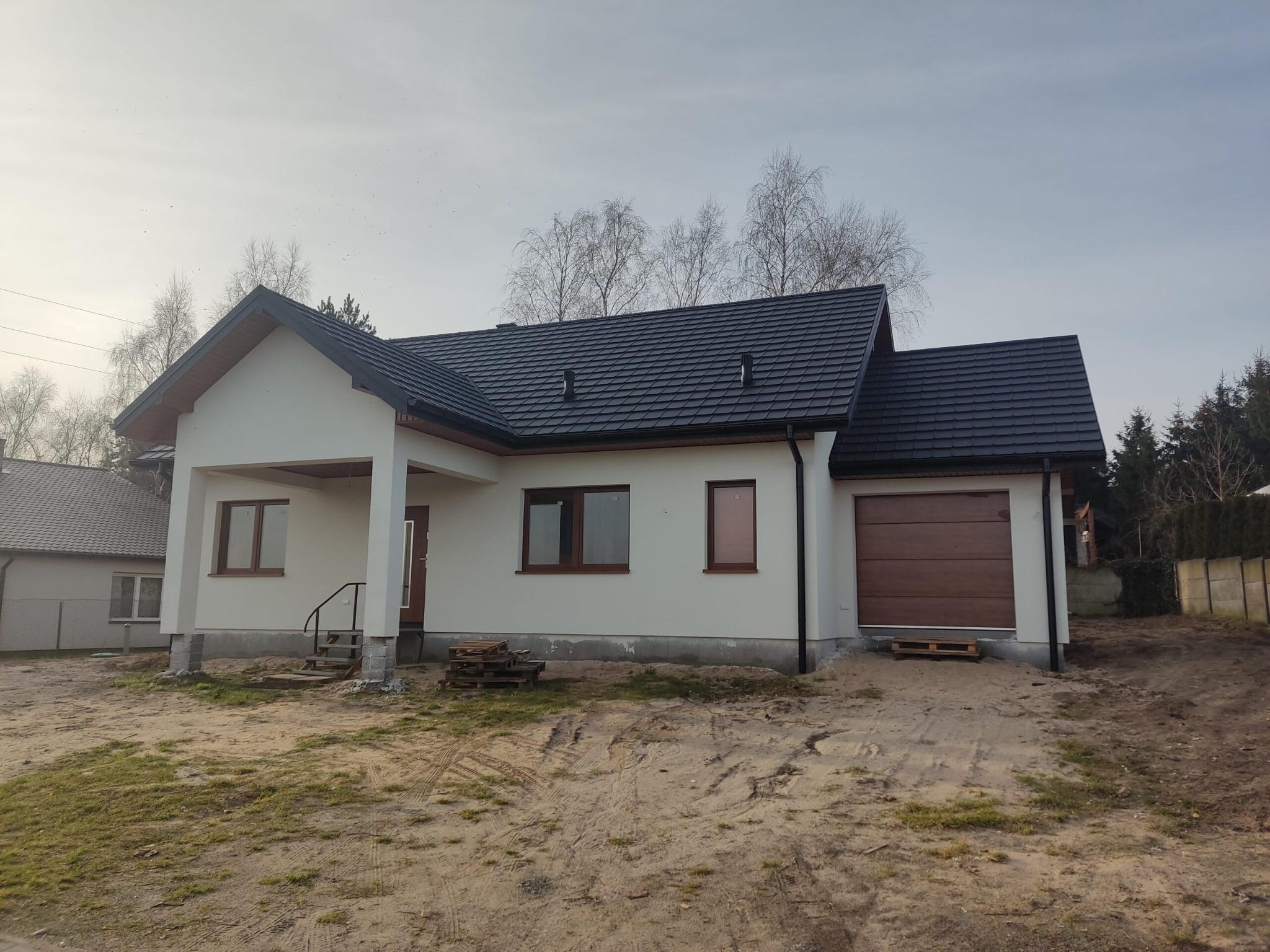 Niedokończony parterowy dom z garażem, biała elewacja, ciemna dachówka, teren wokół domu w trakcie zagospodarowywania, widoczne palety budowlane.
