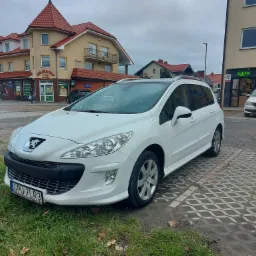 Biały Peugeot 308 SW zaparkowany na ażurowym parkingu przed budynkiem z Żabką i Delikatesami Pod Wieżą.