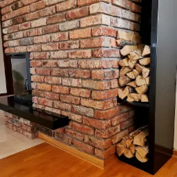 Fragment ściany z cegły klinkierowej z wbudowanym kominkiem i półką na drewno, widoczne drewno kominkowe.