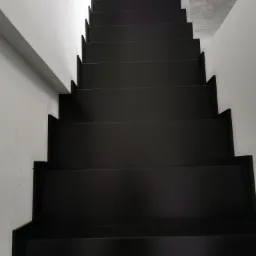 Nowoczesne, czarne schody z minimalistycznym wykończeniem, widok z g&oacute;ry na stopnie, białe ściany po bokach, szara podłoga na g&oacute;rze.