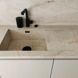 Beżowy blat kamienny z wbudowanym zlewem i czarną armaturą, widoczne białe szafki kuchenne pod spodem.