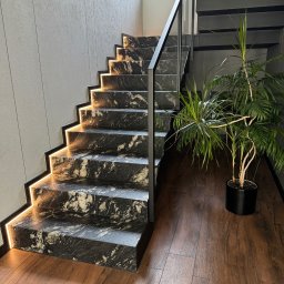 Kamieniarstwo Pietra Naturale - Schody z czarnego kamienia z białymi żyłami, podświetlone ciepłym światłem LED, z metalową balustradą. W tle widoczna zieleń roślin doniczkowych.