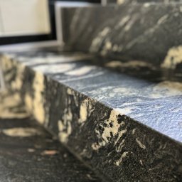 Kamieniarstwo Pietra Naturale - Schody z ciemnego kamienia z unikalnym wzorem, widoczne z bliska, z delikatnym rozmyciem tła. Krawędź stopnia oświetlona, podkreślająca fakturę kamienia.