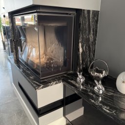 Kamieniarstwo Pietra Naturale - Nowoczesny kominek z blatem z czarnego kamienia o wyraźnym użyleniu, zdobiony srebrnymi figurkami zajączków i dekoracyjną misą. Szare tło i jasna podłoga.