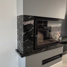 Kamieniarstwo Pietra Naturale - Nowoczesny kominek z obudową z czarnego, wzorzystego kamienia, z widocznym paleniskiem i elementami dekoracyjnymi wewnątrz. Szare ściany i podłoga.