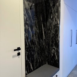 Kamieniarstwo Pietra Naturale - Wnęka z siedziskiem i czarnym, wzorzystym kamieniem na ścianie, uzupełniona o wieszaki. Minimalistyczny design, białe szafki i szuflady, czarne uchwyty.