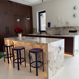 Kamieniarstwo Pietra Naturale - Nowoczesna kuchnia z wyspą wykończoną blatem z unikalnego kamienia z podświetleniem LED. Granatowe hokery dodają elegancji. Na blacie stoi doniczka z czerwoną poinsecją.