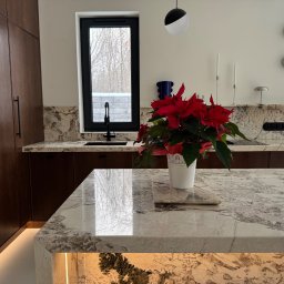 Kamieniarstwo Pietra Naturale - Kuchnia z wyspą i blatem z jasnego kamienia o wyraźnym wzorze. Na blacie stoi biała doniczka z czerwoną gwiazdą betlejemską. W tle okno i zabudowa kuchenna.